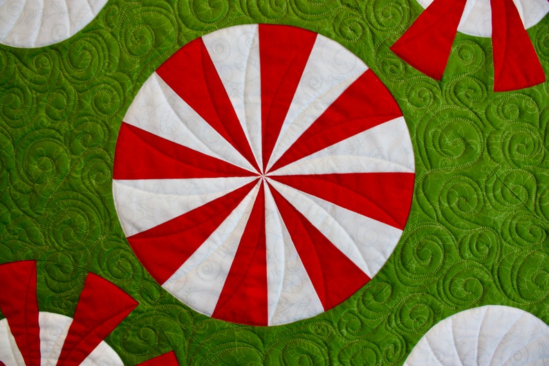 May include: Close-up of a quilted fabric with a repeating pattern of red and white peppermint candies on a green background. The candies have a spiral design. The green background has a swirl pattern.