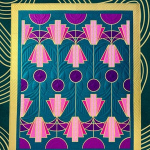 May include: A teal and gold quilt pattern with a geometric design featuring pink and purple shapes. The finished size is 145 cm x 175 cm. The quilt pattern is by Jennifer Wagner.