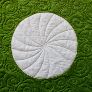 May include: Close-up of a quilted textile with a white circular design in the center. The circle has a spiral pattern. The background is a vibrant green with a swirling pattern, and the stitching is visible.
