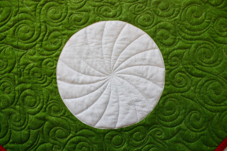 May include: Close-up of a quilted textile with a white circular design in the center. The circle has a spiral pattern. The background is a vibrant green with a swirling pattern, and the stitching is visible.