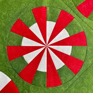 May include: Close-up of a Christmas-themed quilt with a repeating pattern. The design features red and white peppermint-like circles set against a green background with a swirling pattern. The quilt is made of fabric.