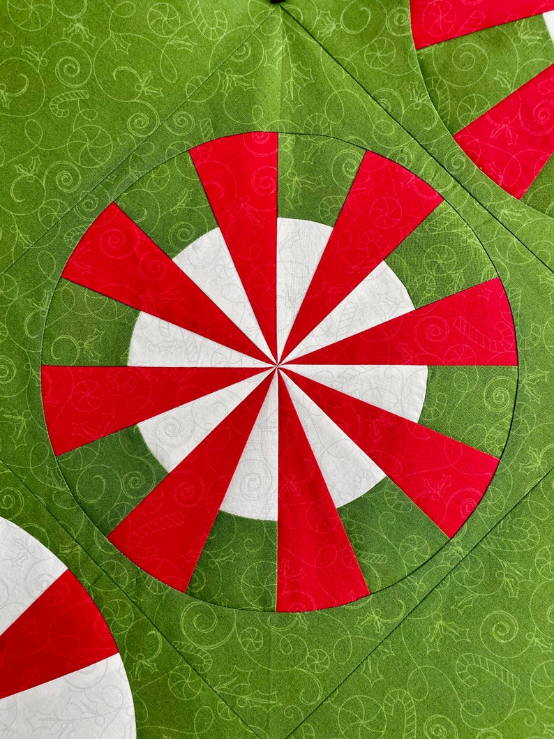 May include: Close-up of a Christmas-themed quilt with a repeating pattern. The design features red and white peppermint-like circles set against a green background with a swirling pattern. The quilt is made of fabric.