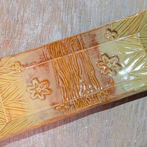 May include: Rectangular ceramic serving dish with a yellow and brown glaze. The dish features embossed floral and geometric patterns. The center has a textured, wood-like design. A decorative piece for serving food.