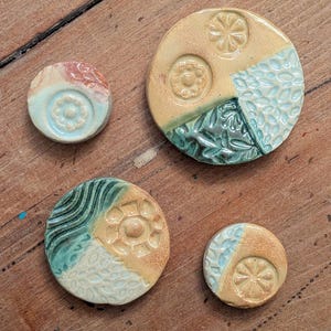 May include: Four handmade ceramic pieces in various sizes and designs. The pieces feature a mix of colors including beige, teal, and coral. Each piece has a unique pattern, including floral and geometric designs. The pieces are arranged on a wooden surface.