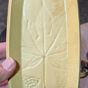 May include: A rectangular ceramic tray in a muted yellow hue featuring a central embossed maple leaf design. The surface displays a textured pattern along the corners and edges, with a smooth, slightly raised border framing the interior area.