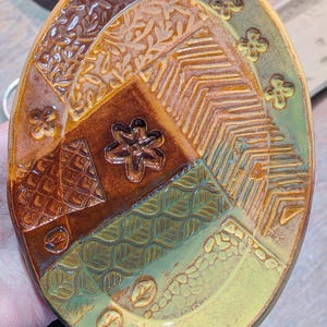 May include: Handmade ceramic oval dish with a patchwork design. The dish features various textured patterns in shades of brown, green, and yellow. Decorative floral and geometric motifs are incorporated throughout the design.