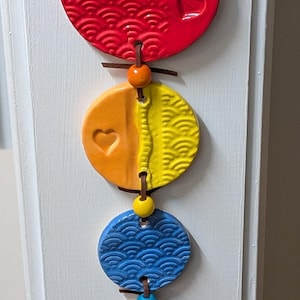 May include: A hanging ceramic ornament featuring five colorful, textured, circular pieces. The top red piece has heart and wave patterns. The other pieces are orange, yellow, blue, green, and brown, each with unique designs and connected by leather cords.