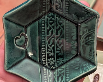 May include: A hexagonal ceramic dish with a glossy dark teal glaze. The surface features a central panel with intricate embossed vine and floral patterns, flanked by smooth sections. One side of the dish displays a small, raised heart-shaped detail.