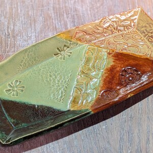 May include: A rectangular ceramic serving dish with a unique design. The dish is divided into sections of green, brown, and tan, each with embossed floral and geometric patterns. The edges are slightly raised, and the overall aesthetic is rustic.