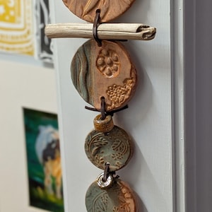 May include: A handcrafted ceramic wall hanging featuring several circular, textured pieces in shades of brown and green. The pieces are connected by leather cords and a small wooden dowel. The top piece has a floral design.