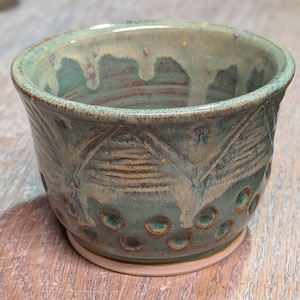 May include: A handmade ceramic bowl with a teal glaze and textured design. The bowl features a series of circular cutouts near the base and a decorative pattern around the middle. The rim has a dripped glaze effect.