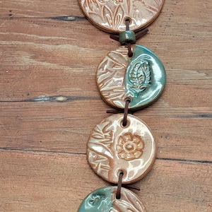 May include: A vertical wall hanging featuring five circular ceramic discs connected by a dark brown leather cord. The discs display embossed floral and botanical patterns, finished in a combination of glossy terracotta and teal green glaze.
