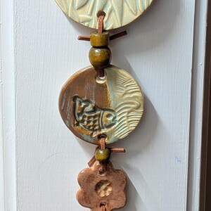 May include: A vertical wall hanging featuring a series of ceramic discs and flower-shaped pieces strung on a brown leather cord. The largest top disc is pale blue with a floral relief, followed by a brown and blue disc with a fish motif.