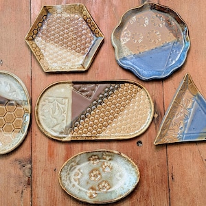 May include: A collection of six ceramic trinket dishes in various shapes including hexagonal, oval, triangular, and scalloped. The dishes feature glazed surfaces in shades of brown, tan, and blue, with embossed textures like honeycombs, paw prints, and floral patterns.