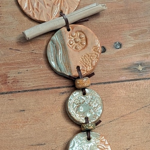 May include: A handcrafted necklace featuring multiple ceramic pendants in shades of peach, green, and cream. The pendants have embossed floral and patterned designs, connected by leather cords. A small metal key charm hangs at the bottom.