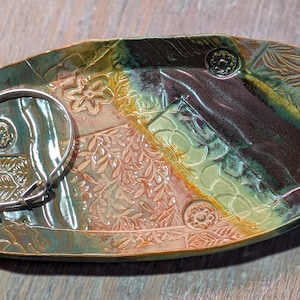 May include: Oval-shaped ceramic trinket dish with a silver bracelet. The dish features a patchwork design with floral and geometric patterns in shades of green, brown, and yellow. Decorative accents include raised circular elements.