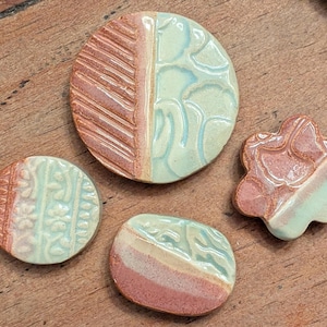 May include: Four handmade ceramic pieces in shades of pink, light blue, and cream. The pieces include two circles, a rounded square, and a flower shape. Each piece has a unique textured design.