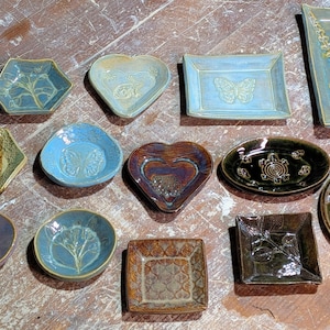 May include: A collection of handcrafted ceramic dishes in various shapes, including oval, heart, and square. The dishes feature embossed designs and a range of colors, such as blue, brown, and green. These decorative pieces are ideal for serving or display.