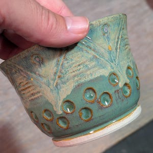 May include: Handmade ceramic bowl with a teal glaze and textured design. The bowl features a series of circular indentations and a white base. The bowl is held by a hand.