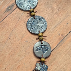 May include: A handcrafted necklace featuring a series of circular, blue-grey ceramic pendants with wave and fish designs. The necklace is strung on a brown cord and accented with wooden beads. The pendant sizes vary.