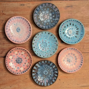 May include: A collection of six circular ceramic bowls arranged in a cluster. Each bowl features a unique reactive glaze pattern with varying color palettes including shades of pink, blue, grey, and cream, displaying intricate radial dot designs.