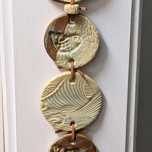 May include: A vertical wall hanging featuring five circular ceramic discs connected by brown leather cords. The discs display embossed patterns of ocean waves and fish in shades of cream, light blue, and brown, with a small dark bead at the bottom.