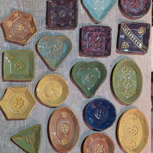 May include: A collection of small, handcrafted ceramic trinket dishes in various shapes including hearts, ovals, hexagons, triangles, and squares. Each dish features unique embossed floral or geometric patterns and a variety of glazed colors.
