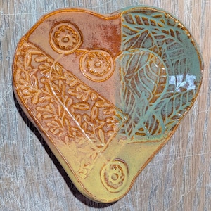 May include: Heart-shaped ceramic dish with a divided design. One side features a floral pattern in shades of orange and yellow, while the other side has a leaf pattern in teal and brown. The dish has a glossy finish.