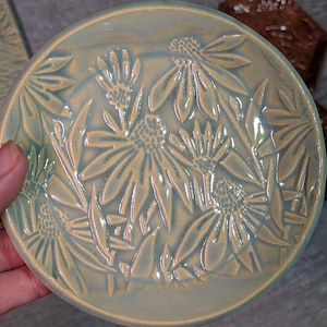 May include: A round ceramic plate featuring a raised, embossed floral pattern of coneflowers. The surface has a glossy glaze with a blend of soft yellow and light blue tones, highlighting the textured petals and centers of the flowers.