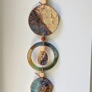 May include: A vertical wall hanging featuring a series of circular ceramic discs and a small pendant connected by a brown leather cord. The ceramic pieces display various embossed floral and geometric patterns in shades of green, blue, and brown.