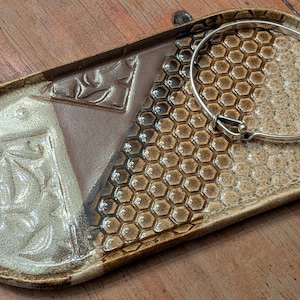 May include: An oval ceramic tray featuring a multi-textured surface with a smooth brown section, a cream-colored area with embossed leaf patterns, and a honeycomb-textured side. A thin, silver-toned metal bangle bracelet rests on the tray.