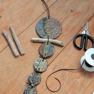 May include: A handcrafted ceramic wind chime with a series of textured, circular elements in shades of green, brown, and gray, strung together with leather cord. The design includes floral and geometric patterns. Driftwood and scissors are also visible.