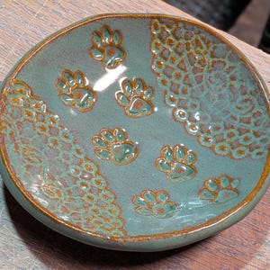 May include: A teal ceramic dish with a golden rim and paw print designs. The dish has a textured, lace-like pattern on one side and several paw prints scattered across the surface. The paw prints and lace pattern are in a golden-brown color.