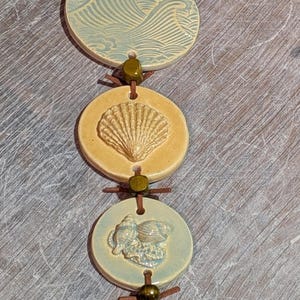 May include: A vertical wall hanging featuring five circular ceramic discs in shades of pale blue and yellow, strung together with brown leather cord and small metallic beads. Each disc displays embossed ocean-themed motifs including waves and shells.