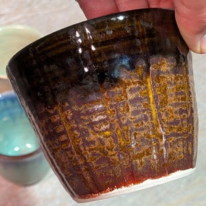 May include: A handmade ceramic cup with a textured surface. The cup features a gradient of colors, transitioning from dark brown at the top to a reddish-brown and gold in the middle, with a white base. The cup is held by a hand.