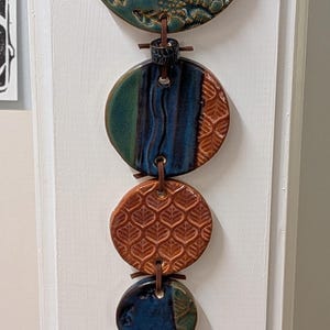 May include: A vertical wall hanging featuring six circular ceramic discs in varying sizes, connected by a brown leather cord. The discs display glazed finishes in shades of teal, blue, and terracotta, with embossed floral and geometric patterns.