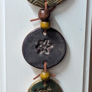May include: A vertical wall hanging featuring five circular ceramic discs in shades of teal, dark brown, and green, strung together with a brown leather cord and interspersed with small wooden beads. The discs display embossed patterns.
