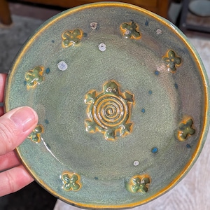 May include: Handmade ceramic plate with a green glaze and a raised turtle design in the center. The plate features embossed flower and dot accents, with a gold-colored rim. A unique, decorative piece for the home.