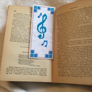 Op de afbeelding: Een close-up van een boek met een blauw en wit borduurwerk-bladwijzer met een solsleutel. Het boek is open op een hoofdstuk met de titel "Hoofdstuk" en de tekst luidt "Food wresht and which...A friend to help find one to hear."