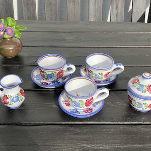 May include: A set of hand-painted ceramic teacups with saucers, a creamer, and a sugar bowl. The white cups and bowls feature a blue rim and are decorated with red and blue floral patterns. A small brown vase with purple flowers is also present.