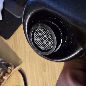 Può includere: Primo piano di un oggetto metallico nero con uno schermo a rete circolare. L'oggetto ha una finitura lucida e sembra far parte di una struttura più grande. Lo schermo a rete è realizzato in un materiale fine color argento.