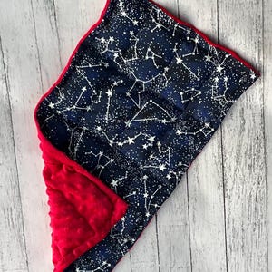 May include: A reversible baby blanket with a navy blue constellation pattern on one side and a red, textured fabric on the other. The blanket has a red border and is designed for comfort and warmth.
