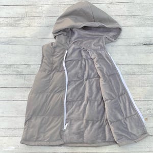 May include: A gray, quilted vest with a hood and a white zipper. The vest is sleeveless and has a soft, plush interior. The exterior is a solid gray color. The hood is attached to the vest with a zipper.