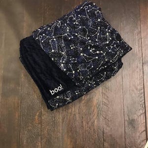 May include: A folded blanket featuring a dark navy blue fabric with a white celestial constellation pattern. One side of the blanket is a solid dark navy blue textured material with the word "boo!" embroidered in white lowercase letters.