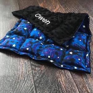 May include: A small, square, quilted blanket with a celestial design. The blanket features a blue and purple galaxy pattern with white stars. The top layer is black and has the name "owen" embroidered in white.