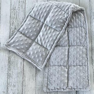 May include: A light gray weighted blanket featuring a soft minky fabric with a raised dimpled texture. The blanket is constructed with a grid of square stitched compartments that create a quilted appearance across the entire surface of the item.