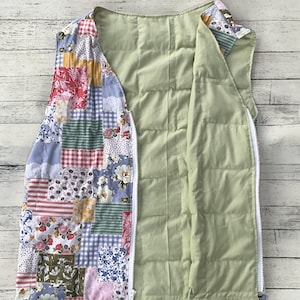 May include: Open, quilted vest with a patchwork design on the exterior. The patchwork features various floral, gingham, and solid fabric squares in shades of red, blue, pink, and green. The interior is a solid light green, and the vest has a white zipper.