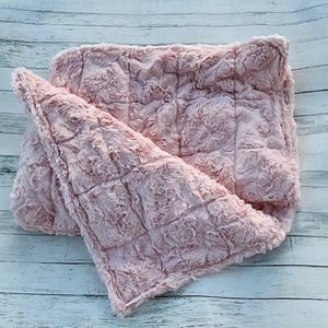 May include: A soft, pink faux fur blanket with a quilted pattern. The blanket is folded over, showing the plush texture and color variations. The blanket is made of a soft, fluffy material.