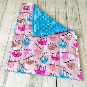 May include: A rectangular baby blanket featuring a light pink background with a repeating pattern of brown, bright pink, and blue sloths hanging from tree branches. The reverse side of the blanket is a solid bright blue fabric with a raised dot texture.
