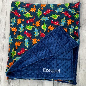May include: A weighted blanket featuring a dark blue background with a repeating pattern of colorful dinosaurs in red, orange, green, and light blue. The reverse side is a solid dark blue minky fabric with a dimpled texture and the name Ezequiel.